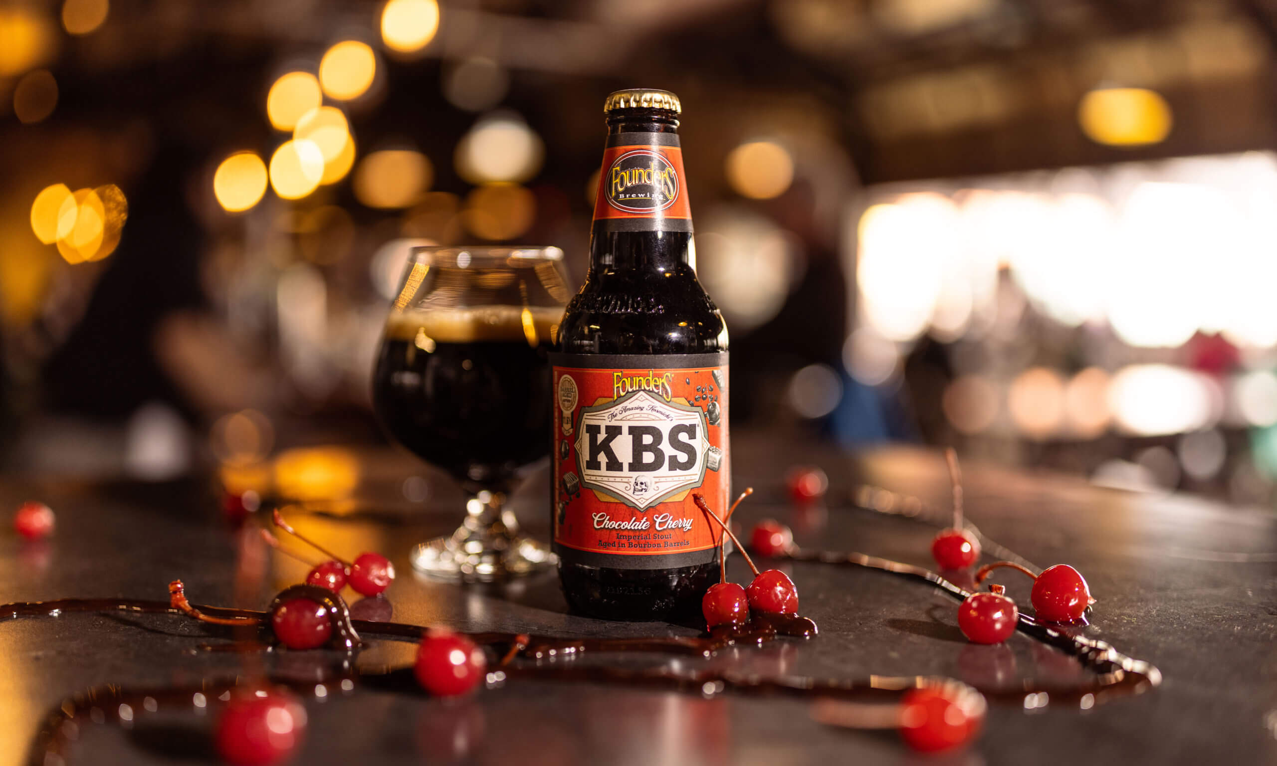 Bottle of KBS Chocolate Cherry and snifter pour on a table surrounded by cherries and chocolate sauce