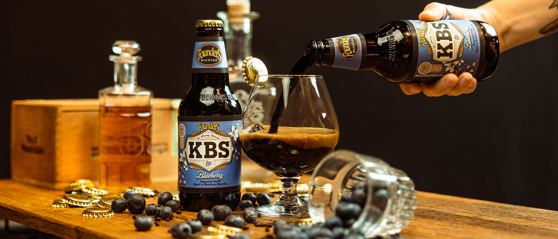 Bottle of KBS Blueberry and snifter pour on table with blueberries and bottle caps