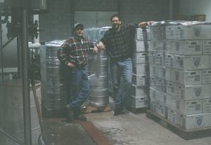 Two men, Dave and Mike, wearing flannel shirts standing by beer kegs and cases of beer