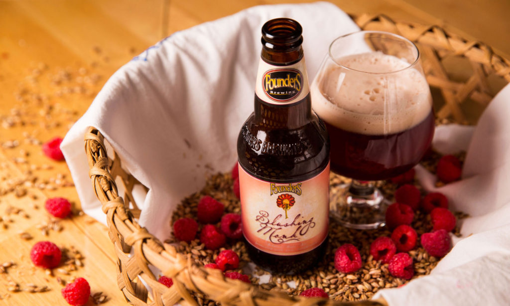 blushing monk bottle next to snifter of blushing monk beer in basket filled with grain and raspberries