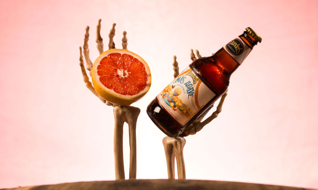 Skeleton hands holding a grapefruit and bottle of Founders Más Agave