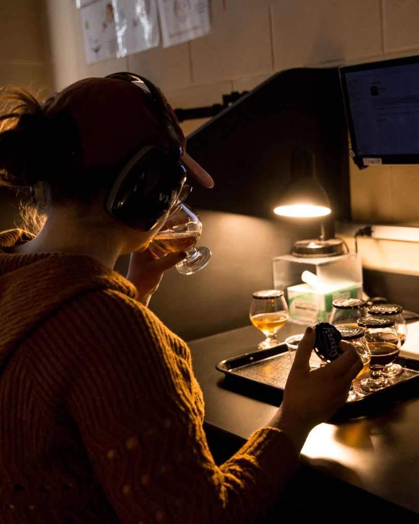 person wearing headphones sipping beer from snifter under dim light in a lab setting