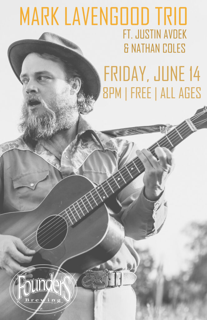 Black and white photo of a man with a beard and hat playing guitar.